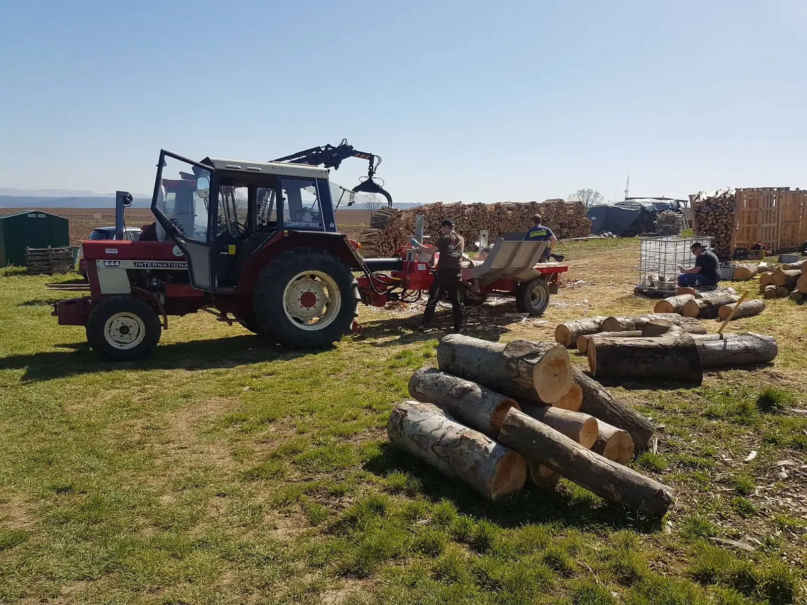 Traktor mit Kran und Spalter, daneben Helfer und Stammholz auf dem Arbeitsplatz